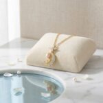 Gold chain necklace with a crystal-encrusted crescent moon pendant and a pink pearl accent, displayed on a cream velvet stand.